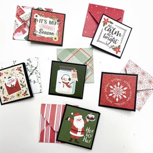 May include: A collection of small, square Christmas cards with matching envelopes. The cards feature festive designs, including a snowman in a jar, Santa Claus, and the phrases "It's My Favorite Season" and "All is Calm, All is Bright."