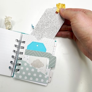 May include: Open spiral-bound notebook with various patterned pages and pockets. A black and white dotted tag is held by a hand. The notebook features a teal tag and a cloud graphic.