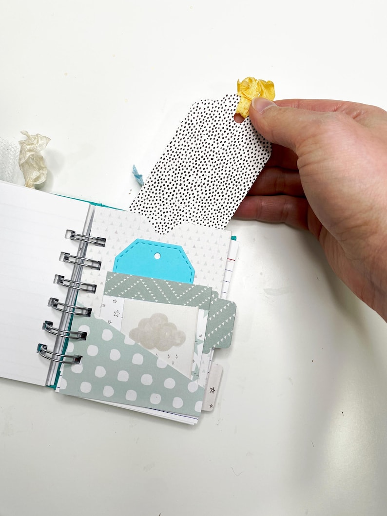 May include: Open spiral-bound notebook with various patterned pages and pockets. A black and white dotted tag is held by a hand. The notebook features a teal tag and a cloud graphic.
