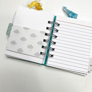 May include: Open notebook with lined pages and a pocket featuring a gray cloud pattern. The notebook has a spiral binding and colorful ribbon bookmarks. The pages are white with blue lines.