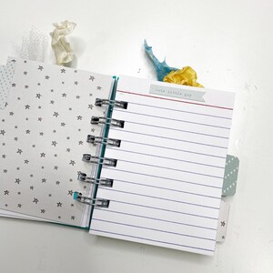 May include: Open spiral-bound notebook with lined pages and a small tag that reads "cute little guy." The notebook has a teal spine and star-patterned pages. Decorative ribbons in white, blue, and yellow are tucked inside.