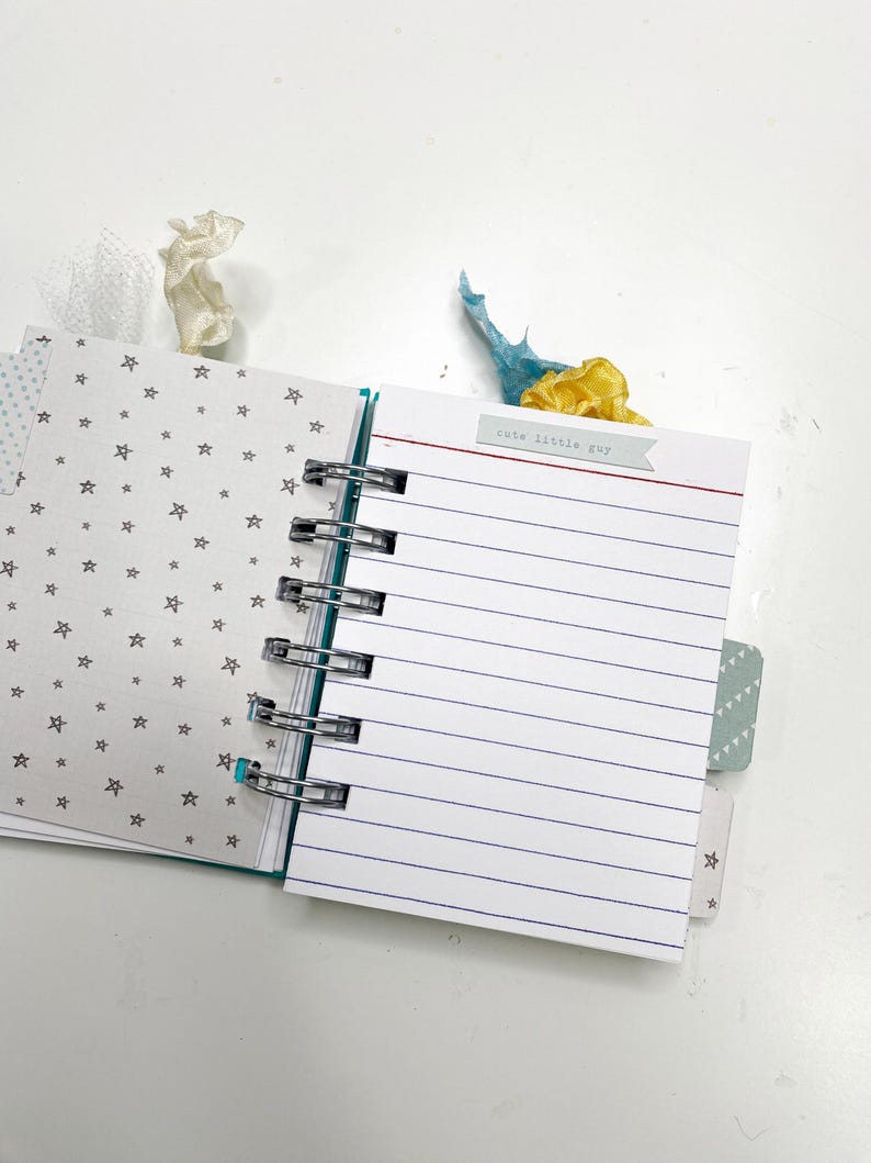 May include: Open spiral-bound notebook with lined pages and a small tag that reads "cute little guy." The notebook has a teal spine and star-patterned pages. Decorative ribbons in white, blue, and yellow are tucked inside.