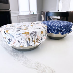 May include: Two white ceramic bowls with fabric covers. One bowl has a blue and white polka dot cover and the other has a white cover with a black and orange print of kitchen utensils and the words "Bake, Blend, Salad, Market Fresh".