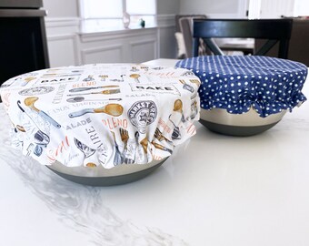 May include: Two white ceramic bowls with fabric covers. One bowl has a blue and white polka dot cover and the other has a white cover with a black and orange print of kitchen utensils and the words "Bake, Blend, Salad, Market Fresh".