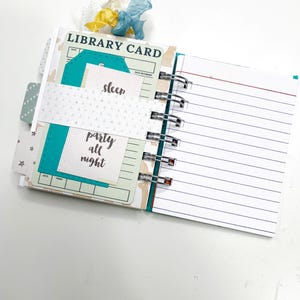 May include: Open journal with lined pages and library card-themed inserts. The inserts are teal and white with the words "sleep" and "party all night." A decorative ribbon in blue, yellow, and white accents the top. The journal is bound with silver metal rings.