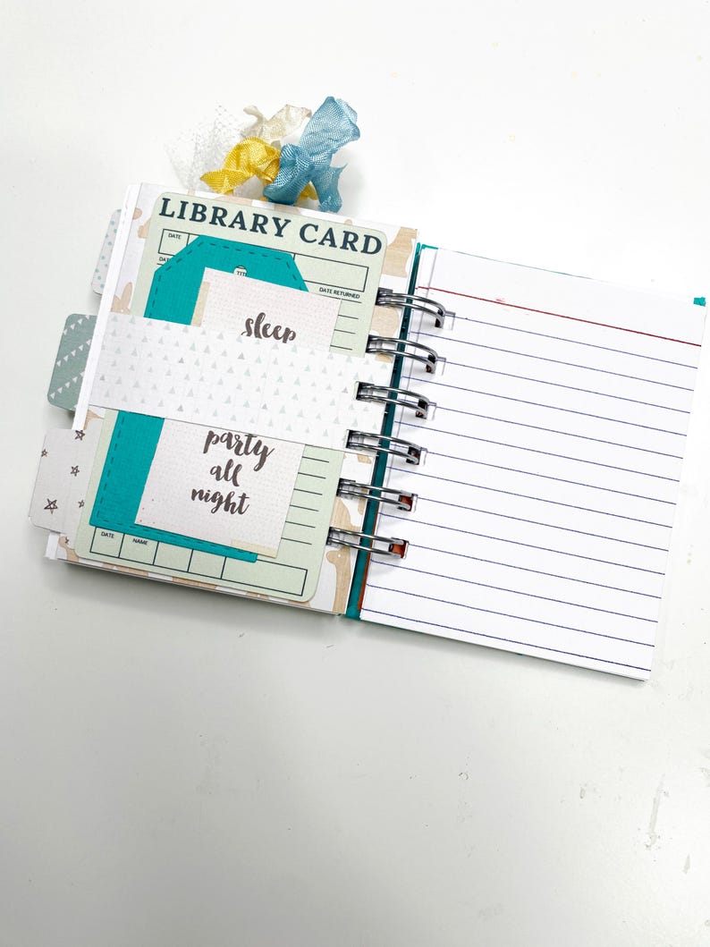 May include: Open journal with lined pages and library card-themed inserts. The inserts are teal and white with the words "sleep" and "party all night." A decorative ribbon in blue, yellow, and white accents the top. The journal is bound with silver metal rings.
