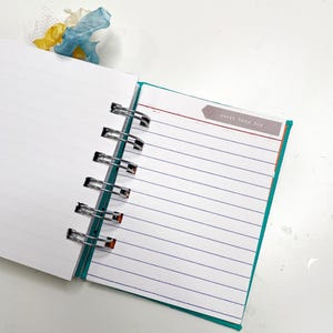 May include: Open notebook with lined pages and a teal border. The text "sweet baby boy" is on a small label. A blue and yellow ribbon bookmark is visible at the top. The notebook is held together with silver metal rings.