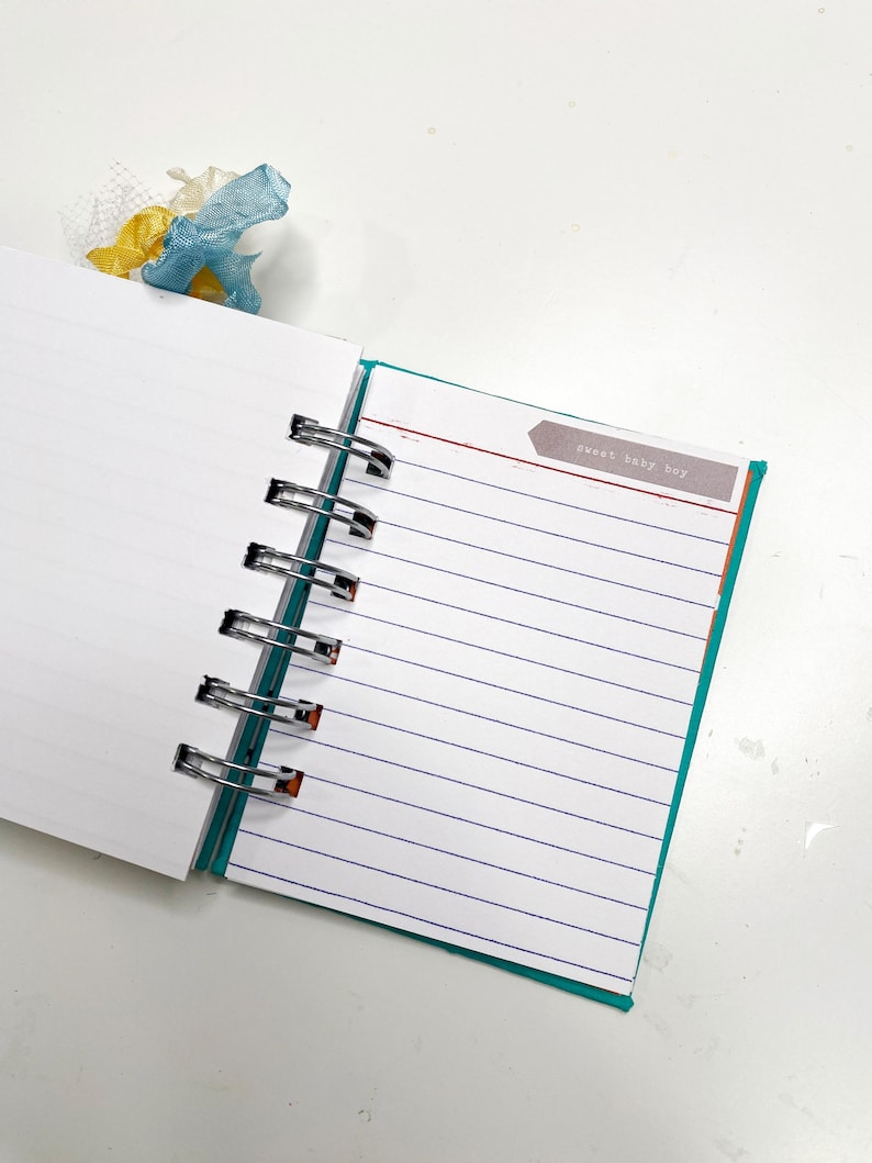 May include: Open notebook with lined pages and a teal border. The text "sweet baby boy" is on a small label. A blue and yellow ribbon bookmark is visible at the top. The notebook is held together with silver metal rings.