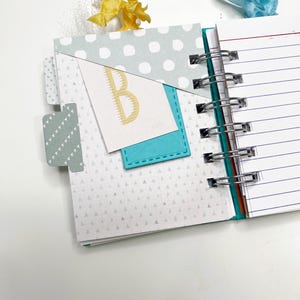 May include: Open notebook with a teal spine and lined pages. The cover features a light blue polka dot pattern and a teal pocket with a card that has a gold letter "B". A yellow ribbon and a blue ribbon are attached to the top.