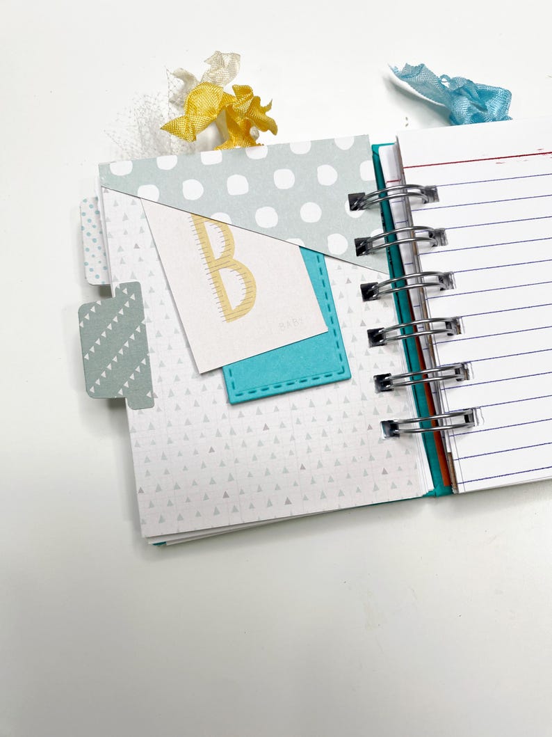 May include: Open notebook with a teal spine and lined pages. The cover features a light blue polka dot pattern and a teal pocket with a card that has a gold letter "B". A yellow ribbon and a blue ribbon are attached to the top.