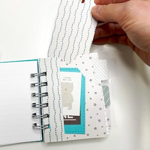May include: A small, open notebook with a spiral binding, featuring a teal cover and interior accents. The pages include a tag with a repeating triangle pattern and a card with a bear illustration and the text "little man cub".