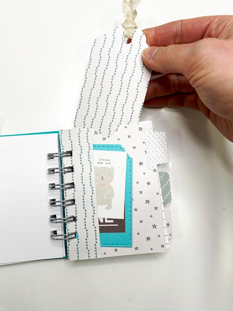 May include: A small, open notebook with a spiral binding, featuring a teal cover and interior accents. The pages include a tag with a repeating triangle pattern and a card with a bear illustration and the text "little man cub".