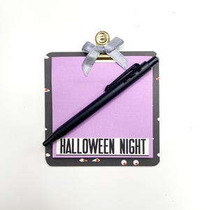 Könnte beinhalten: Ein quadratisches Klemmbrett mit schwarzem Rand und kleinen Augenmotiven, das einen hellvioletten Notizblock mit dem Text HALLOWEEN NIGHT hält. Eine silberne Schleife ist am goldenen Clip befestigt und ein schwarzer Stift liegt diagonal auf dem Block.
