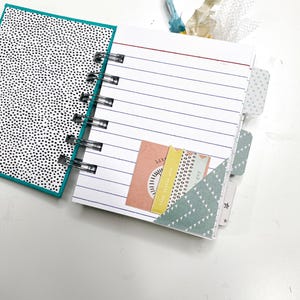 May include: Open notebook with lined pages and a black and white polka dot cover. The notebook has a spiral binding and decorative tabs. A pocket holds colorful paper tags with text and designs.