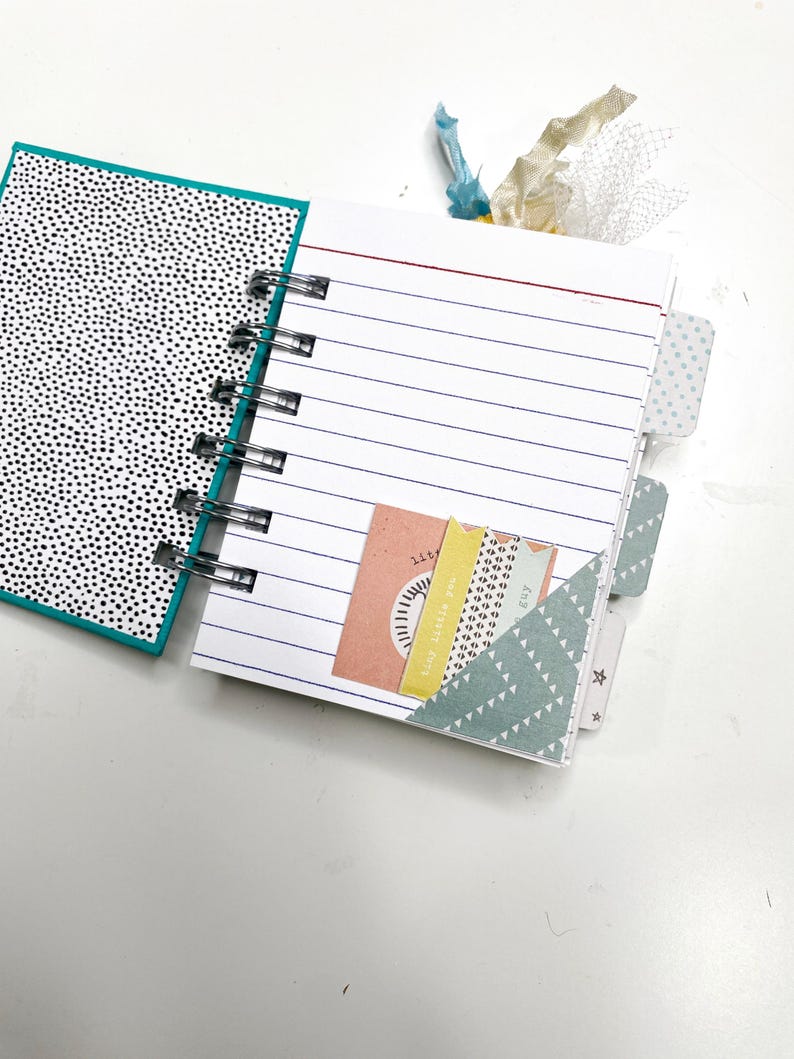 May include: Open notebook with lined pages and a black and white polka dot cover. The notebook has a spiral binding and decorative tabs. A pocket holds colorful paper tags with text and designs.