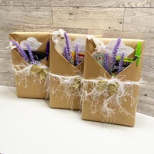 May include: Three brown paper gift boxes, each adorned with a gold glitter bow and white fringe. Each box contains purple lavender sprigs and a pen. The boxes are arranged on a white surface against a wood-paneled wall.