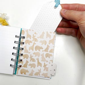 May include: A small spiral-bound notebook with lined pages and a decorative animal print. The cover features a light brown pattern of bears, giraffes, and rabbits. A white tag with a blue ribbon is held near the notebook.