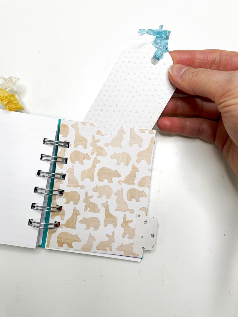 May include: A small spiral-bound notebook with lined pages and a decorative animal print. The cover features a light brown pattern of bears, giraffes, and rabbits. A white tag with a blue ribbon is held near the notebook.