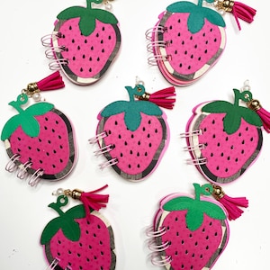 May include: A collection of seven strawberry-shaped mini notebooks. Each notebook features a pink strawberry design with black seeds, a green leaf, and a pink tassel. The notebooks are bound with silver wire and have a pearl accent.