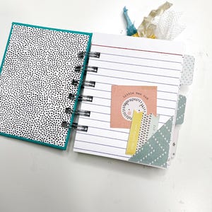 May include: Open spiral-bound notebook with lined pages and decorative tabs. The left page has a black and white polka dot pattern. The right page features a small square with the text "little man cub" and a lion illustration.
