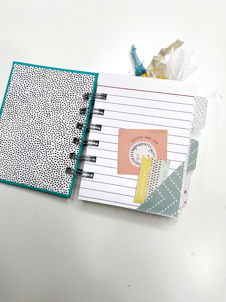 May include: Open spiral-bound notebook with lined pages and decorative tabs. The left page has a black and white polka dot pattern. The right page features a small square with the text "little man cub" and a lion illustration.