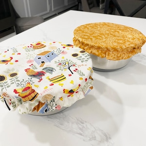 May include: Two fabric bowl covers on a white countertop. One cover is yellow with a white floral pattern. The other cover is white with a colorful rooster and chicken pattern.