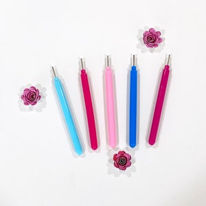 May include: Five paper quilling tools in different colors: pink, blue, light pink, and two shades of red. The tools are arranged in a semi-circle on a white background.