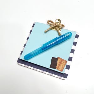 May include: A small square clipboard with a navy and white striped border, holding a stack of light blue paper. A bright blue glittery pen rests on the paper, which features a small graphic of a chocolate bar and a graham cracker. A gold clip with twine.