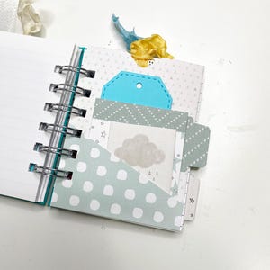 May include: Open spiral-bound notebook with lined pages. The notebook has decorative pages with polka dots, triangles, and a cloud graphic. A blue tag and yellow ribbon are visible. The notebook is open, revealing the interior pages.