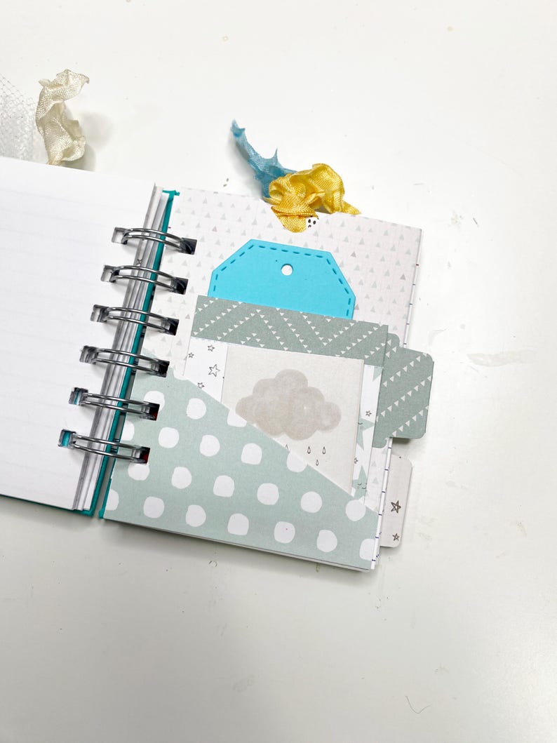 May include: Open spiral-bound notebook with lined pages. The notebook has decorative pages with polka dots, triangles, and a cloud graphic. A blue tag and yellow ribbon are visible. The notebook is open, revealing the interior pages.