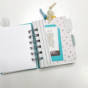 May include: Open spiral-bound journal with blank white pages. The journal has a teal cover and a metal spiral binding. The right side of the journal has decorative pages with star and triangle patterns. A tag with a bear illustration and the text "little man cub" is attached.