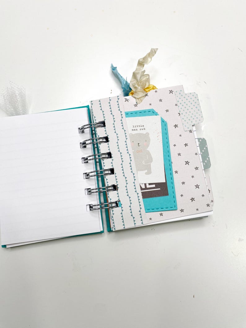 May include: Open spiral-bound journal with blank white pages. The journal has a teal cover and a metal spiral binding. The right side of the journal has decorative pages with star and triangle patterns. A tag with a bear illustration and the text "little man cub" is attached.
