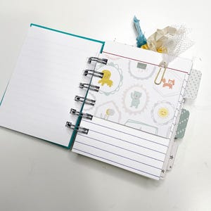 May include: Open spiral-bound notebook with lined pages and a teal cover. The notebook features decorative pages with animal illustrations, a gold paper clip, and a ribbon bookmark. The pages are white with a teal border.