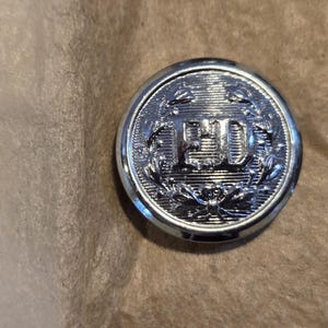 May include: A silver button with a raised, textured design. The button features a wreath of leaves surrounding the letters "FD" in a raised, textured font.