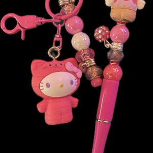 May include: A pink pen with a Hello Kitty charm and a matching keyring. The pen has a gold tip and is adorned with beads. The keyring features a Hello Kitty figure in a pink outfit. The background is black.