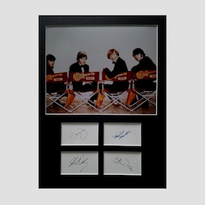 Op de afbeelding: Een ingelijste zwart-witfoto van de leden van de band The Monkees die op regisseursstoelen zitten. De stoelen hebben de naam van de band en de naam van elk lid erop. De foto is gesigneerd door alle vier leden van de band.