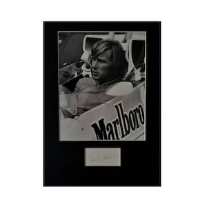May include: Black and white photo of a race car driver in a racing car with a Marlboro logo on the side. The driver is wearing a racing suit and helmet. The photo is signed "James Hunt".