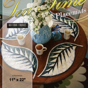 Puede incluir: Manteles individuales con forma de hoja con un diseño blanco y verde, dispuestos sobre una mesa redonda de madera. La imagen incluye un jarrón con flores blancas, tazas de té y una botella decorativa. El texto dice "Tea Time Placemats" y miden 28 cm x 56 cm.