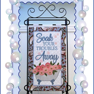 Könnte beinhalten: Ein blauer und weißer gesteppter Wandbehang mit dem Text "Soak Your Troubles Away" und einer weißen Badewanne mit rosa Blumen. Der Behang befindet sich auf einem schwarzen Metallständer. Ein Design von Janine Babich.