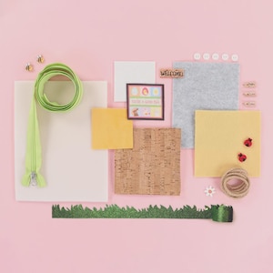 May include: A flat lay arrangement on a pink background features craft supplies. Items include a green zipper, cork squares, felt squares, a framed picture with Easter eggs, and decorative elements like ladybugs and bees.