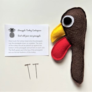 May include: A brown felt turkey head with a yellow beak and red wattle. Two silver pins are included. Instructions are provided on a white card that reads "Pineapple Turkey Centerpiece Just add your own pineapple" and explains how to pin the turkey head onto a pineapple.