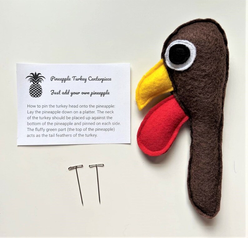 May include: A brown felt turkey head with a yellow beak and red wattle. Two silver pins are included. Instructions are provided on a white card that reads "Pineapple Turkey Centerpiece Just add your own pineapple" and explains how to pin the turkey head onto a pineapple.
