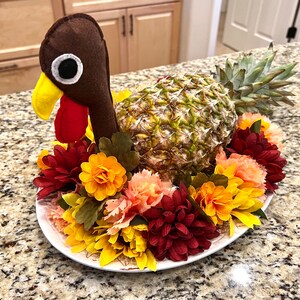 May include: A brown felt turkey with a red beak and a pineapple for a body, surrounded by a wreath of yellow, red, and orange flowers.