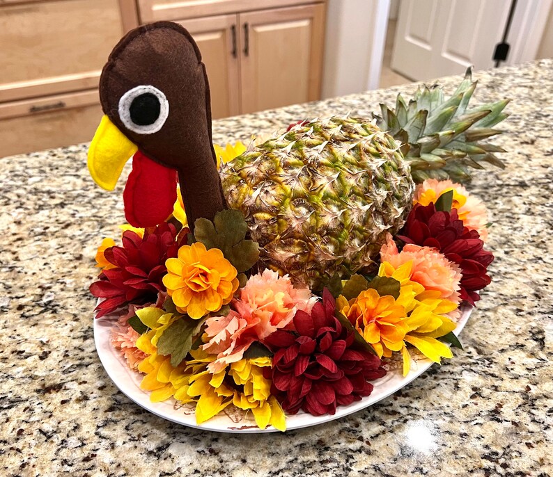 May include: A brown felt turkey with a red beak and a pineapple for a body, surrounded by a wreath of yellow, red, and orange flowers.