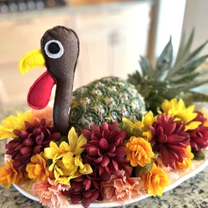 May include: A felt turkey figurine sits atop a pineapple, surrounded by a ring of burgundy, yellow, and peach flowers. The pineapple is on a white plate.