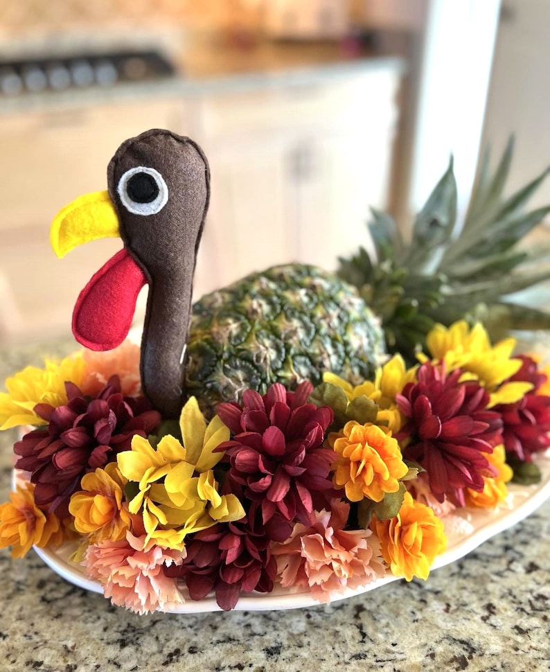 May include: A felt turkey figurine sits atop a pineapple, surrounded by a ring of burgundy, yellow, and peach flowers. The pineapple is on a white plate.
