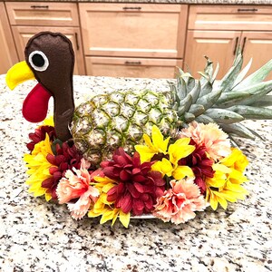 May include: A pineapple with a brown felt turkey head and red felt wattle, surrounded by yellow and red flowers. The pineapple is a Thanksgiving centerpiece.