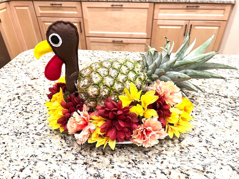 May include: A pineapple with a brown felt turkey head and red felt wattle, surrounded by yellow and red flowers. The pineapple is a Thanksgiving centerpiece.