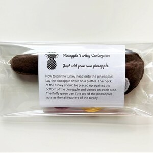 May include: A brown felt turkey head with a white felt beak and a red felt wattle. The turkey head is packaged in a clear plastic bag with a white label that reads "Pineapple Turkey Centerpiece" and "Just add your own pineapple". Instructions for pinning the turkey head onto a pineapple are included on the label.