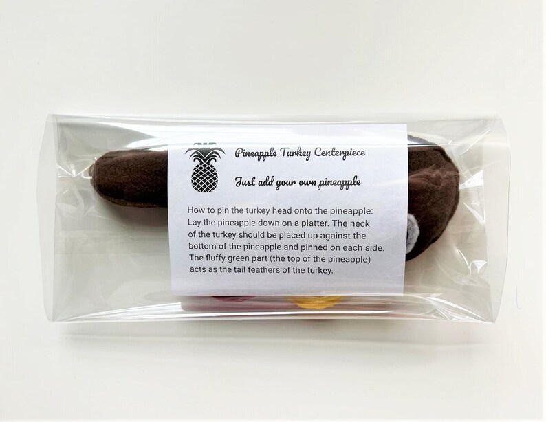 May include: A brown felt turkey head with a white felt beak and a red felt wattle. The turkey head is packaged in a clear plastic bag with a white label that reads "Pineapple Turkey Centerpiece" and "Just add your own pineapple". Instructions for pinning the turkey head onto a pineapple are included on the label.